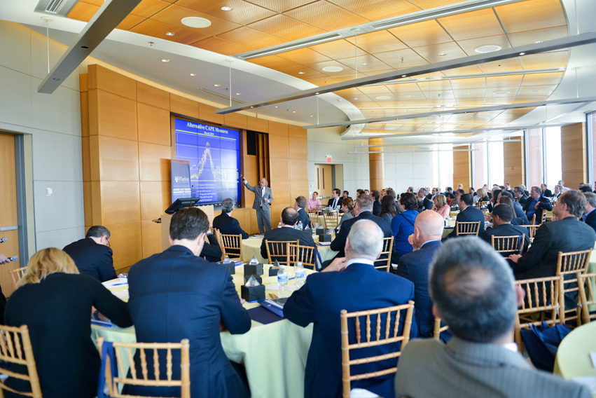 A presenter speaking to an audience in a conference room. The room is filled with attendees seated at round tables. A large screen displays a graph behind the presenter.