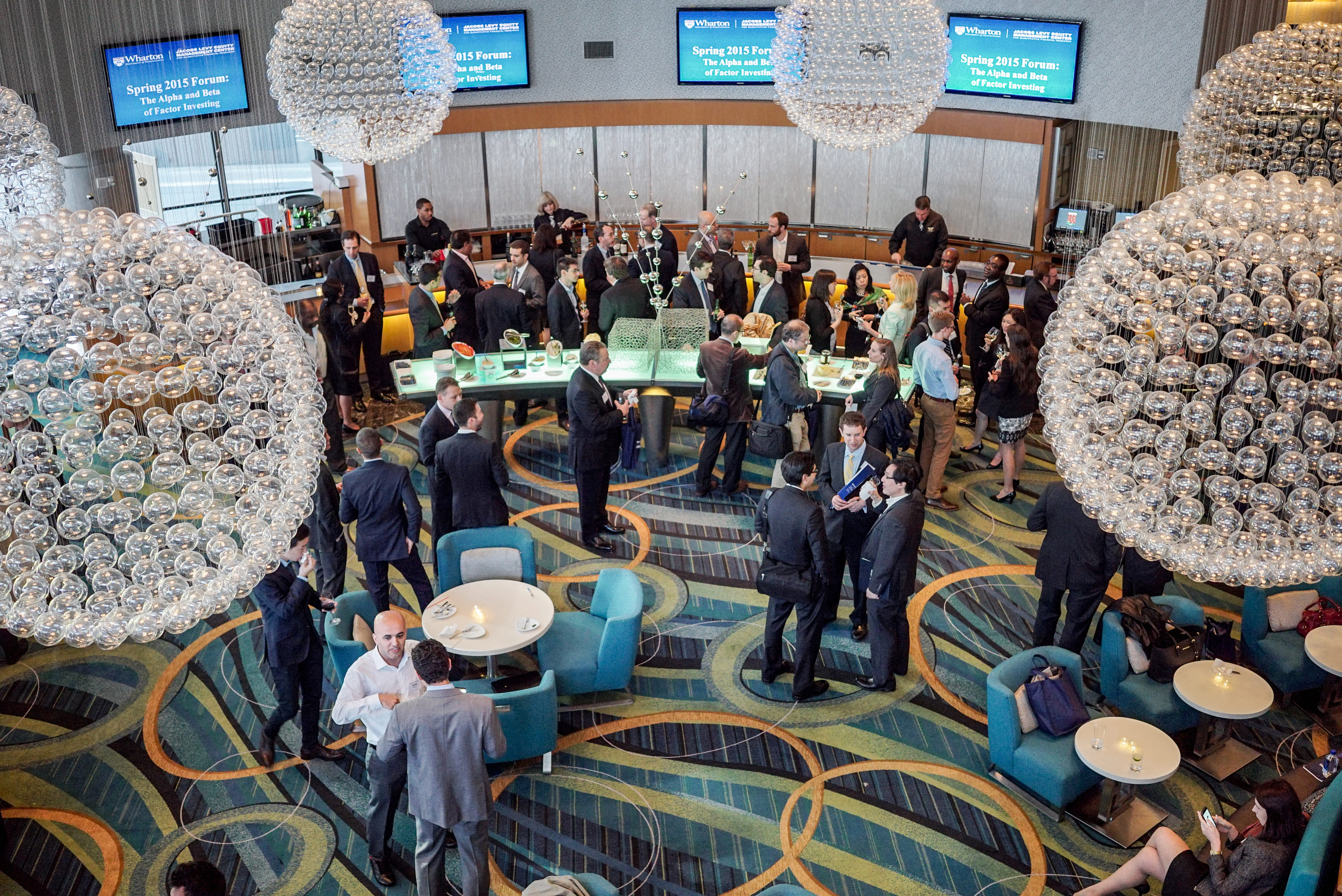 A networking event with participants in formal attire is depicted. The venue features large, spherical chandeliers and modern decor, including a bar area and social seating.