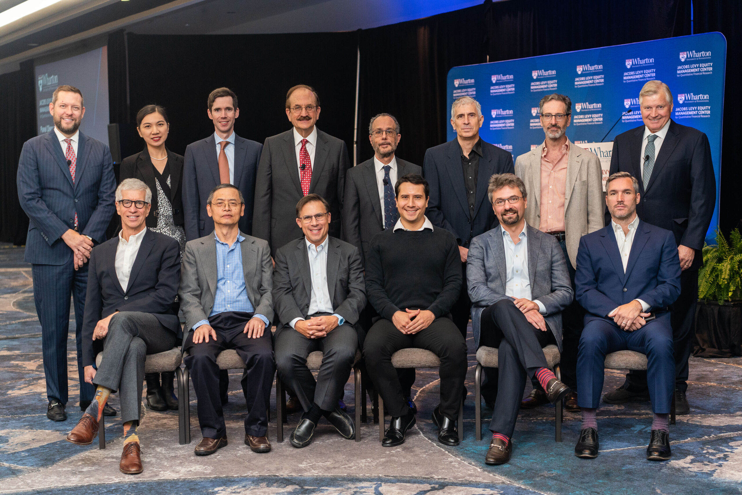 A group of thirteen people posing for a photo, most wearing formal business attire. They are indoors with a backdrop featuring Wharton branding.