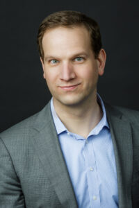 Headshot of a person with short hair, wearing a gray suit and a light blue shirt, set against a dark background.