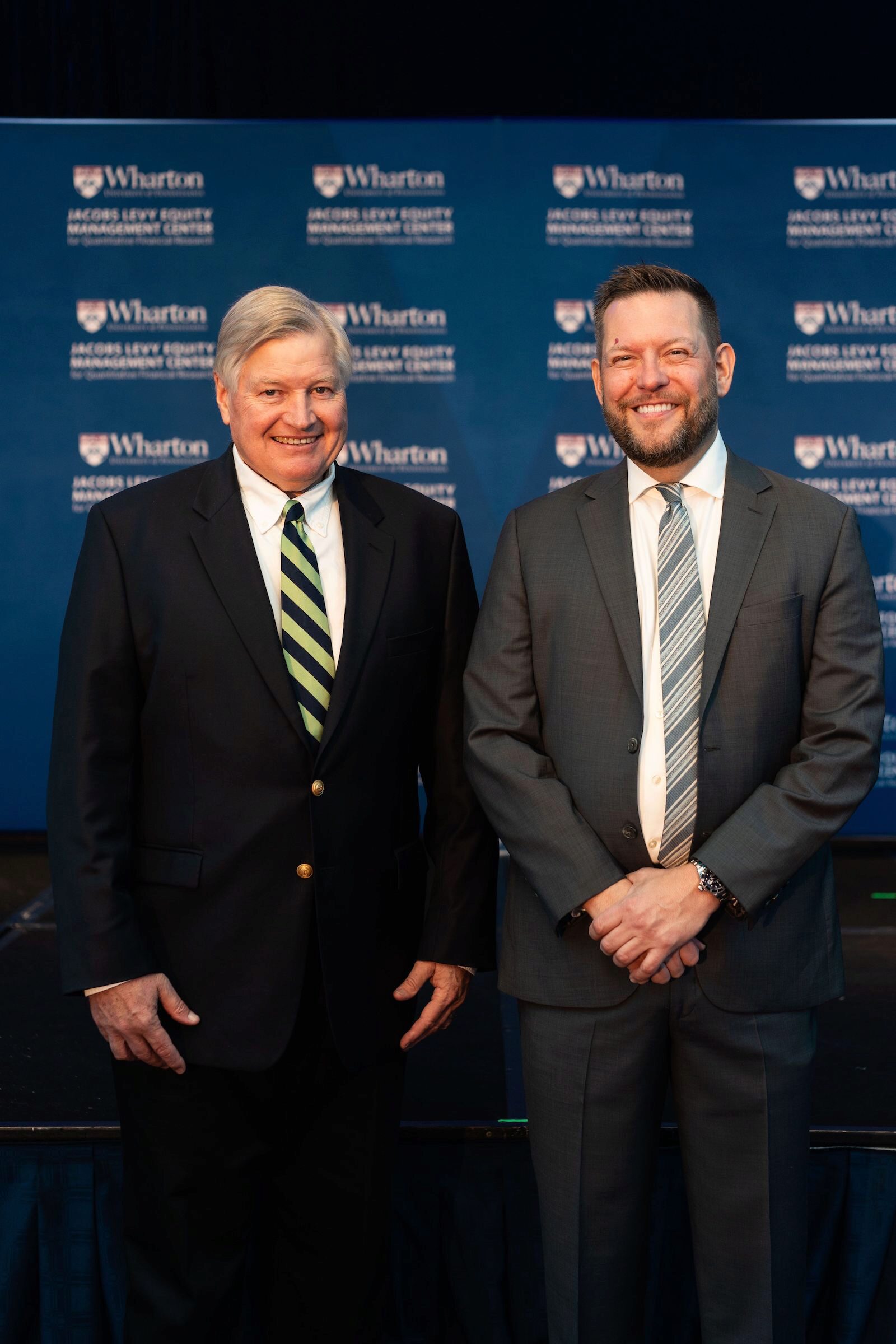 Craig MacKinlay and Christopher Geczy smiling before a blue Wharton background