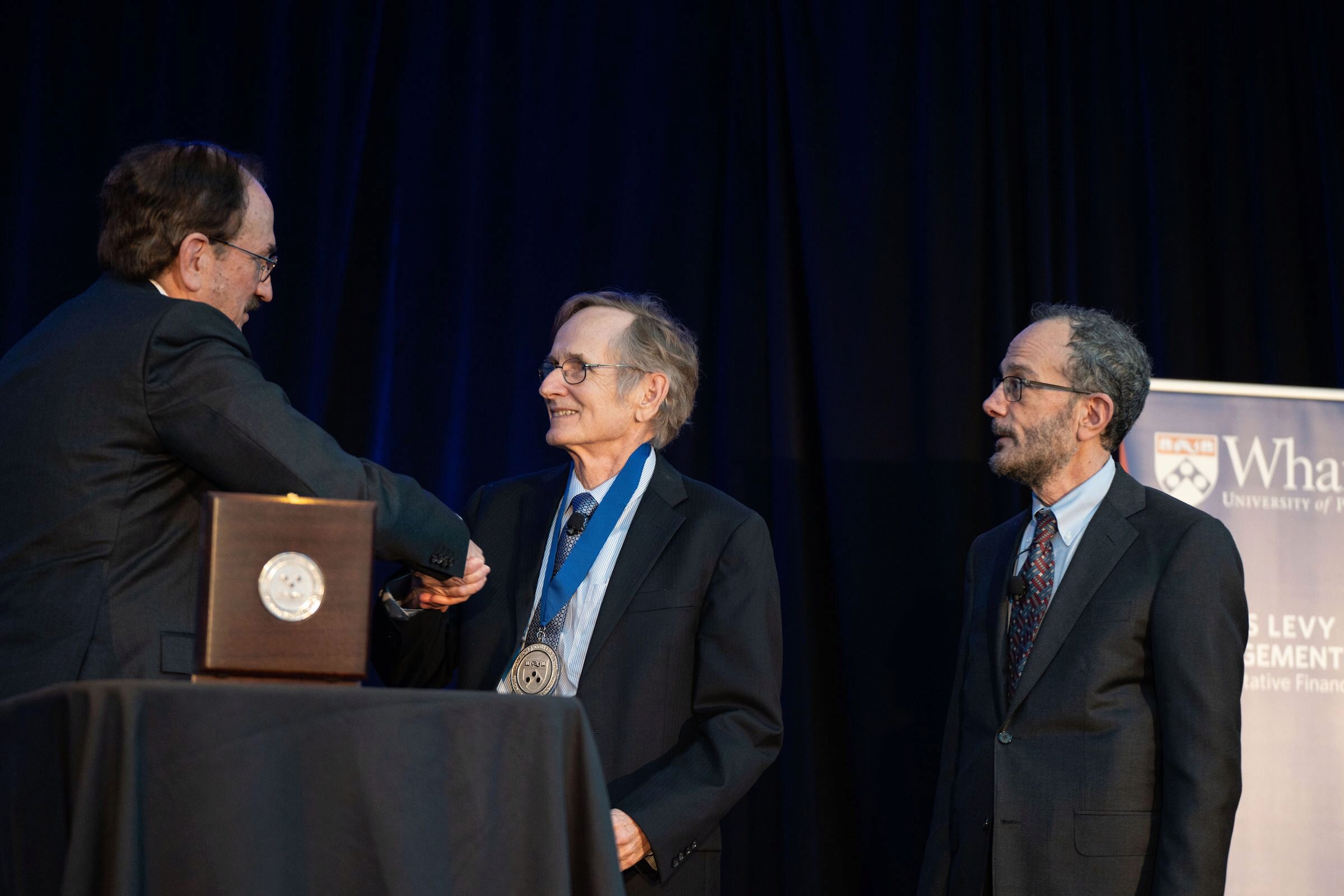 Bruce Jacobs and Albert S. “Pete” Kyle shake hands. Pete wears a medal. Ken Levy stands by.