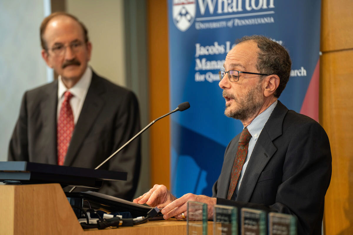 Ken Levy speaks at a podium as Bruce Jacobs watches, during the Frontiers in Quantitative Finance Conference