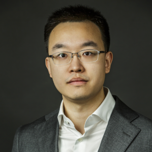 A headshot of a person in glasses before a gray background.