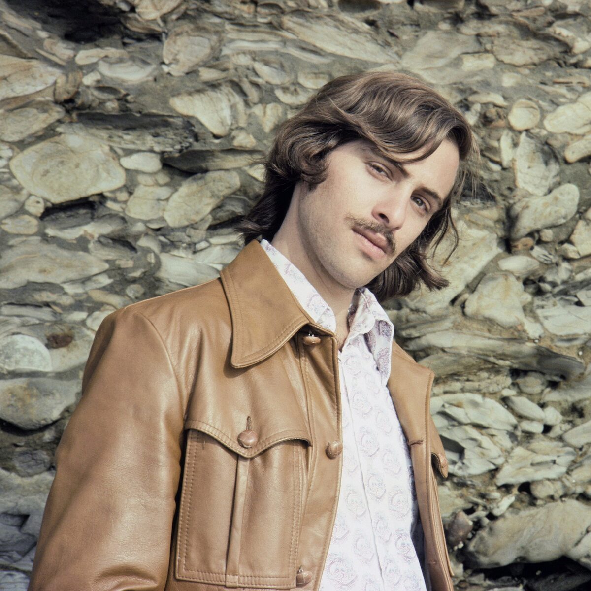 A person in a leather jacket leans against a stone wall