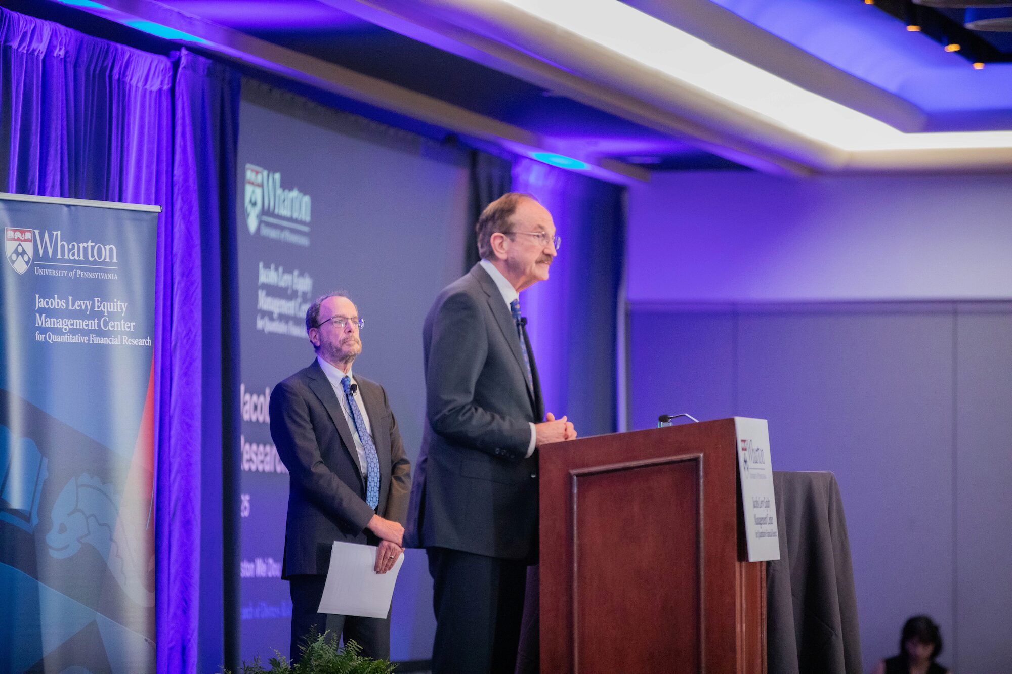A person speaks from a podium while another person watches behind holding papers