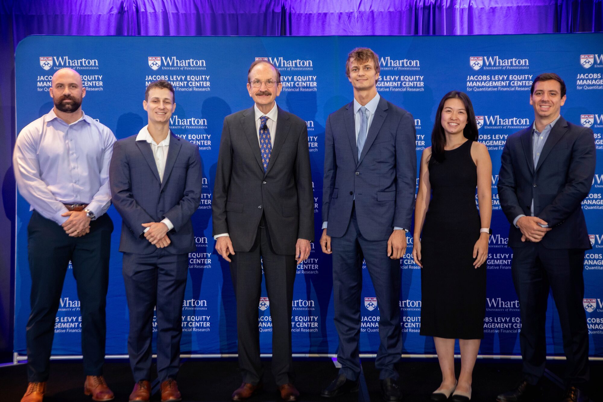 Six people pose for a group photo before a blue banner