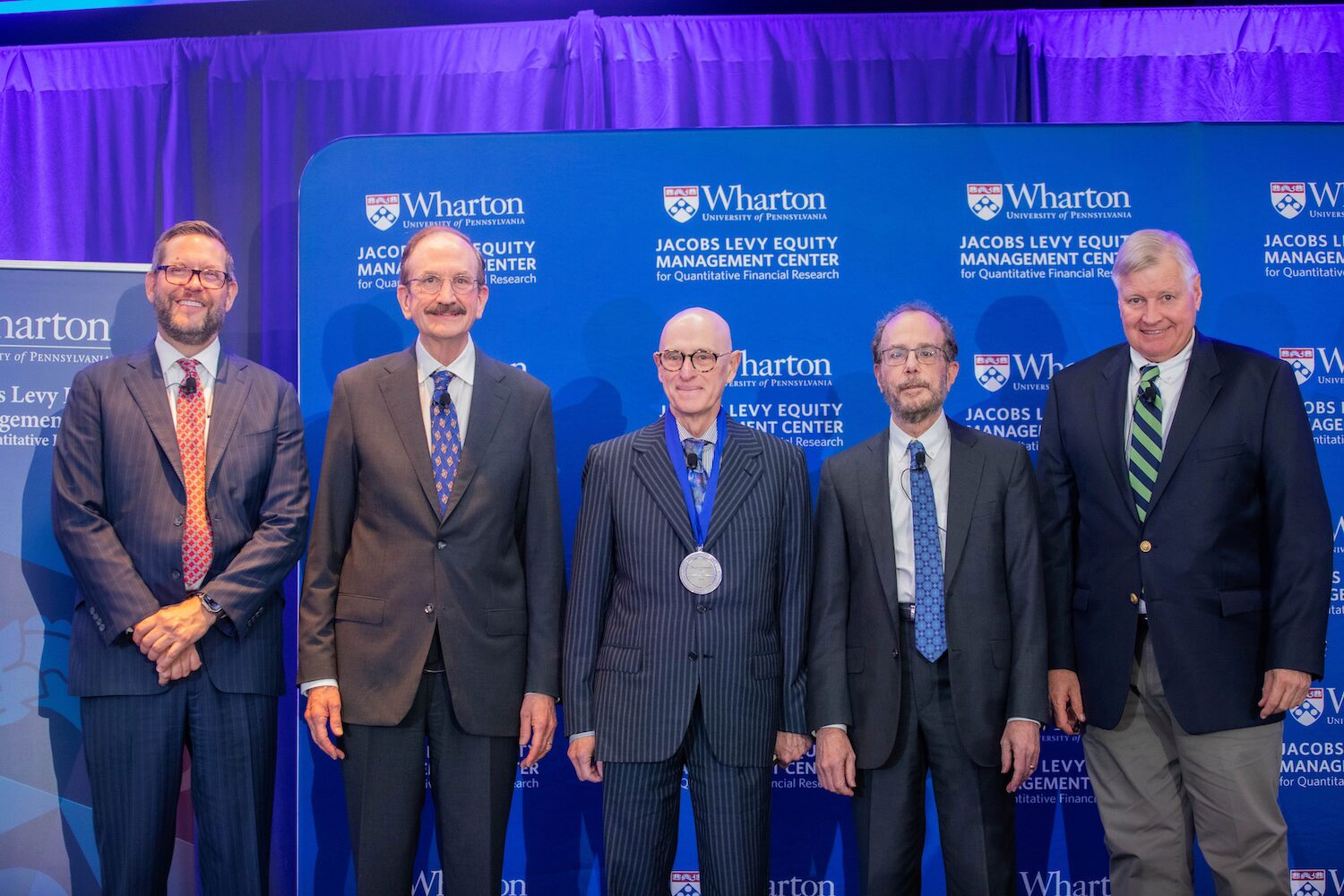 Five people pose for a group photos. The person in the middle wears a medal.
